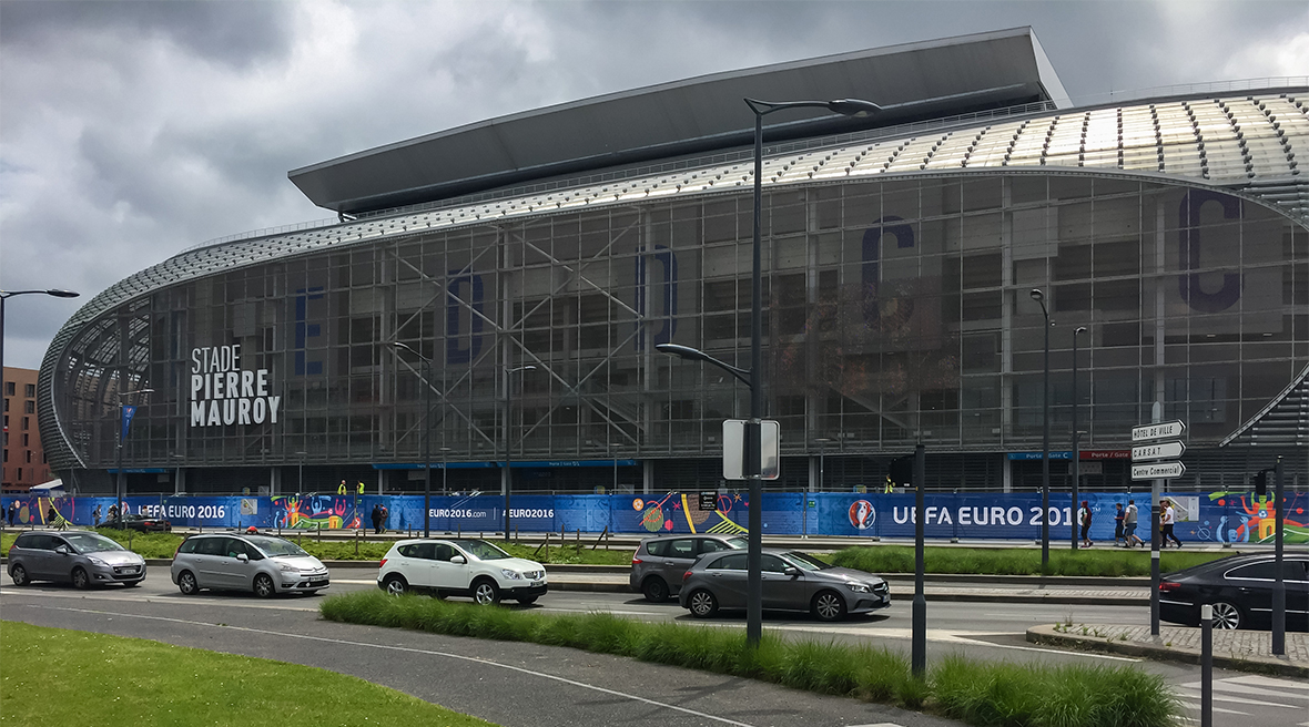 Cars drive past a modern sports stadium, with its name ‘Stade Pierre Mauroy’ prominent, and UEFA Euro 2016 hoardings at ground level